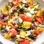 Close up of an in focus serving spoon lifting a scoop of Greek chickpea salad, with more salad out of focus in a serving bowl behind it.