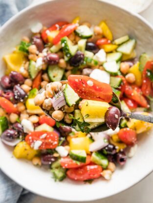 Close up of an in focus serving spoon lifting a scoop of Greek chickpea salad, with more salad out of focus in a serving bowl behind it.