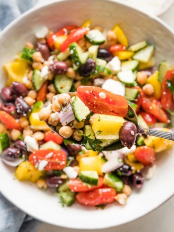 Close up of an in focus serving spoon lifting a scoop of Greek chickpea salad, with more salad out of focus in a serving bowl behind it.