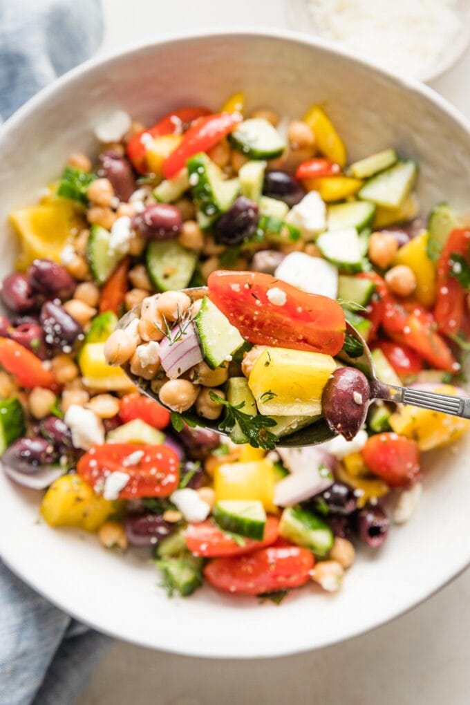 Close up of an in focus serving spoon lifting a scoop of Greek chickpea salad, with more salad out of focus in a serving bowl behind it.