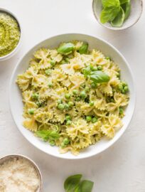 Bowl of pesto pasta and peas surrounded by extra Parmesan, extra pesto, and basil leaves.