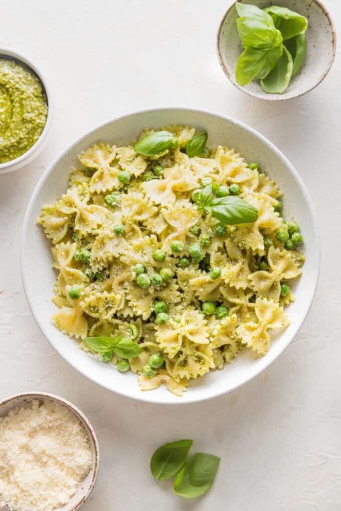 Bowl of pesto pasta and peas surrounded by extra Parmesan, extra pesto, and basil leaves.