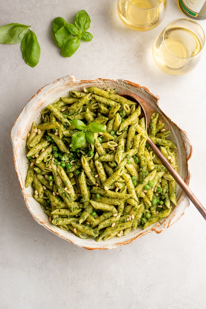 Large serving bowl filled with fresh pesto pasta with peas, surrounded by fresh basil leaves and glasses of white wine.