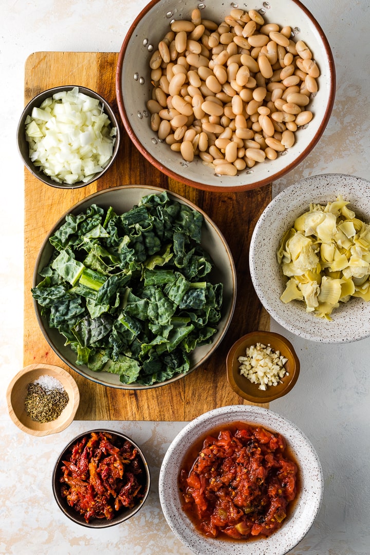 Bowls filled with the ingredients: white beans, kale, onions, artichoke hearts, garlic, diced tomatoes, sun-dried tomatoes, seasoning.
