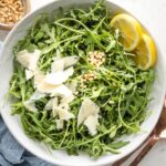 Bowl of arugula salad with shaved Parmesan, lemon slices, and pine nuts.