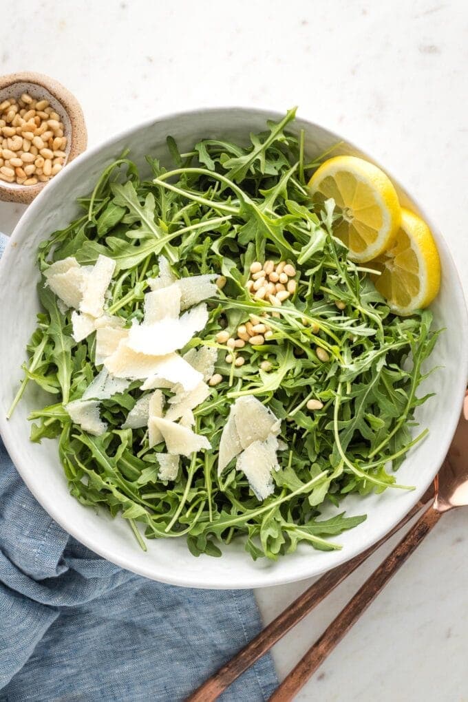 Bowl of arugula salad with shaved Parmesan, lemon slices, and pine nuts.