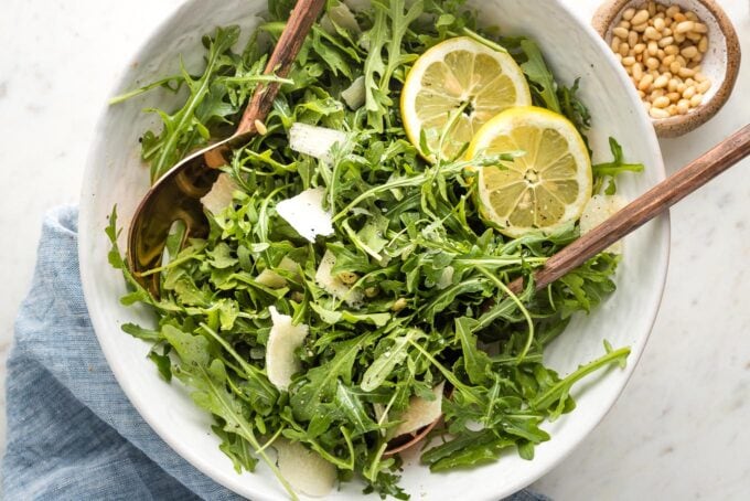 Bowl of arugula salad with shaved Parmesan, lemon slices, and pine nuts.