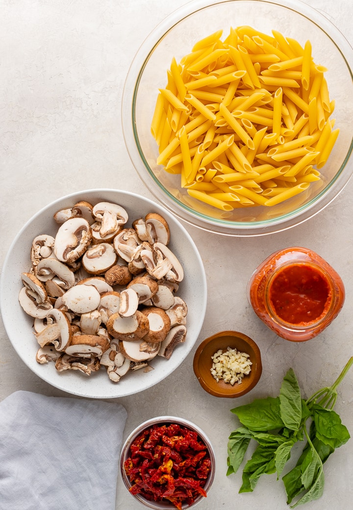 Bowls filled with dried pasta, sliced mushrooms, sun-dried tomatoes, and garlic, plus vodka sauce and fresh basil leaves.