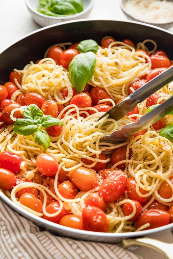 Close up of tongs serving spaghetti with roasted cherry tomatoes straight out of a pan.
