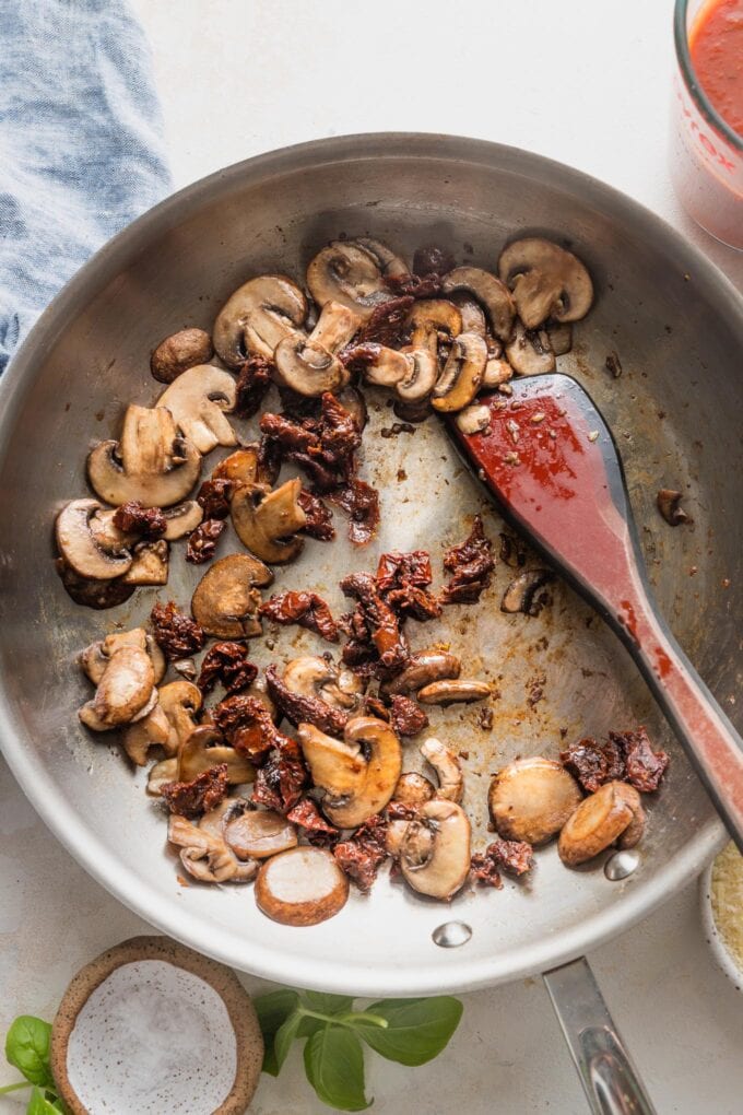 Minced garlic and sun-dried tomatoes added to browned mushrooms in a skillet.