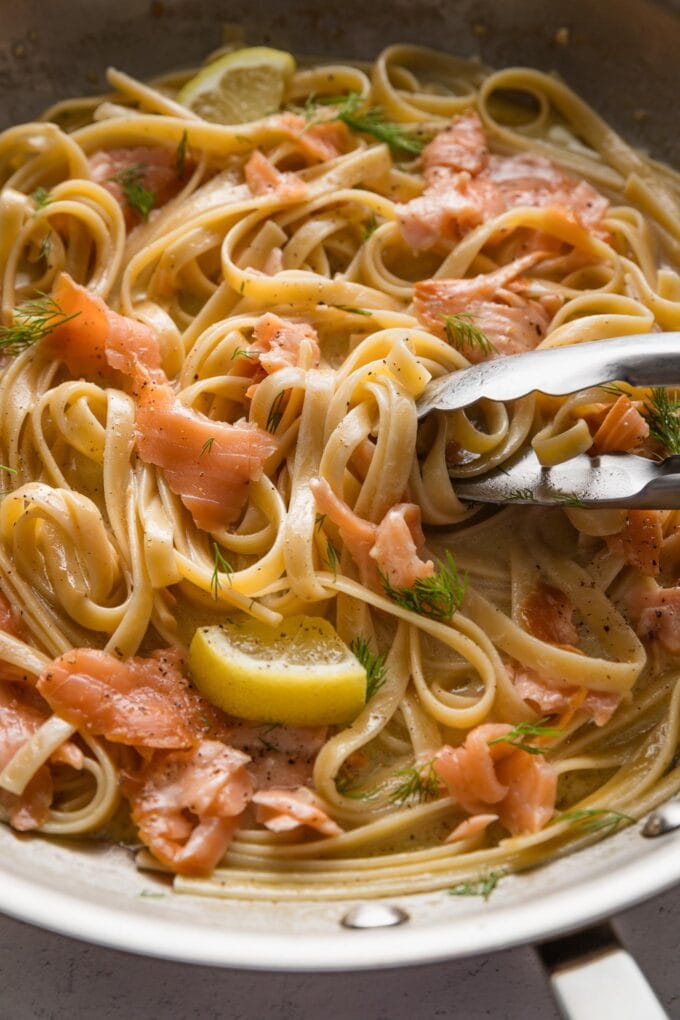 Close up of kitchen tongs twirling and mixing a skillet full of linguine with lemon, cream sauce, and smoked salmon.