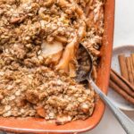 Spoon lifting a serving of old fashioned apple crisp from a brown earthenware baking dish.