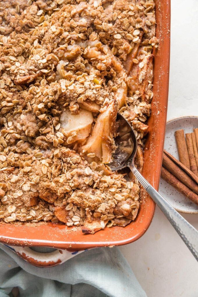 Spoon lifting a serving of old fashioned apple crisp from a brown earthenware baking dish.