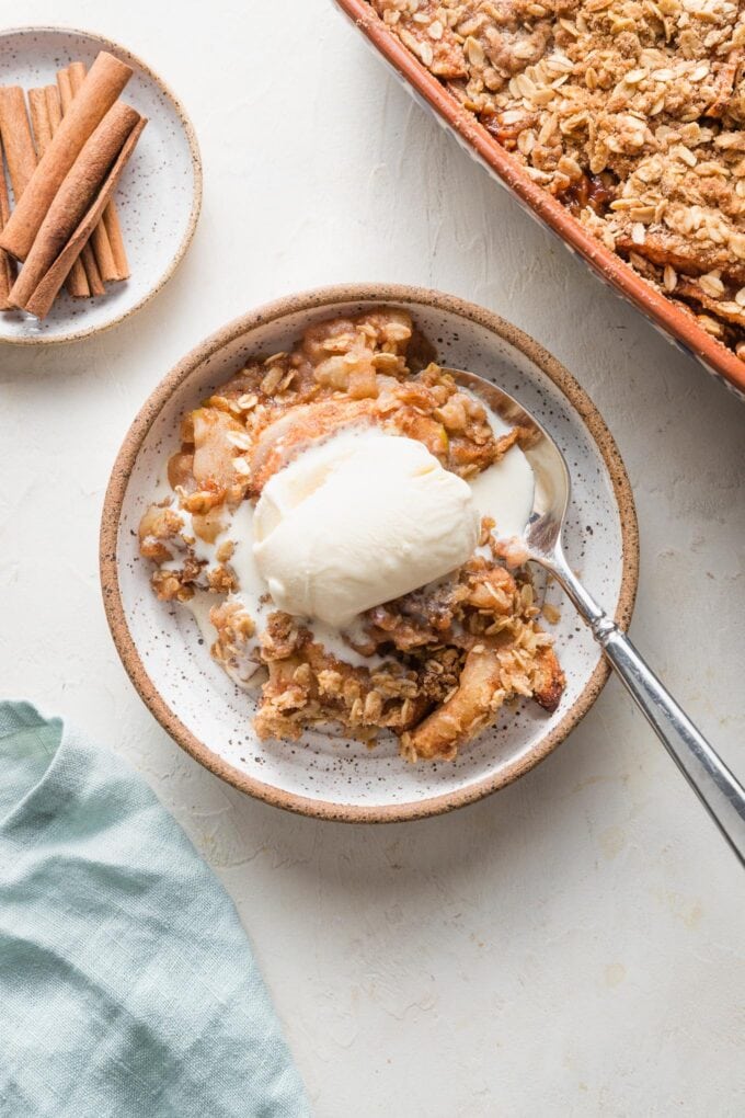 Serving of old fashioned apple crisp with vanilla ice cream in a small dessert bowl, with cinnamon sticks in a bowl nearby.