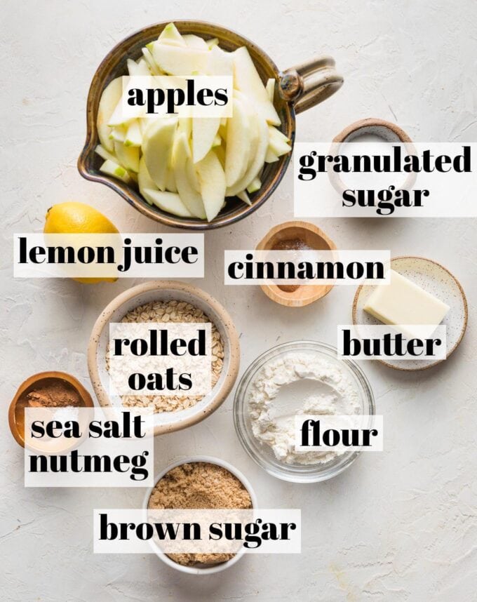 Labeled overhead image of peeled, sliced apples, granulated and brown sugar, flour, rolled oats, a lemon, butter, cinnamon, nutmeg, and sea salt measured and arranged in prep bowls ready to bake.