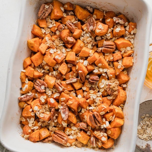 Large baking pan full of chopped sweet potatoes baked in cinnamon honey butter and topped with a light layer of oats, pecans, and brown sugar.