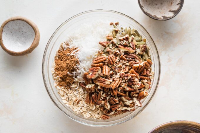 Oats, pecans, coconut flakes, pumpkin seeds, and spices added to a mixing bowl.