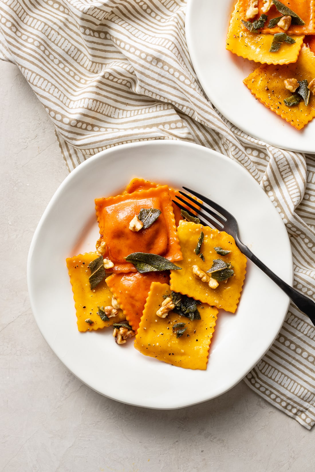 A plate with orange and yellow pumpkin ravioli garnished with sage brown butter sauce and toasted walnuts.