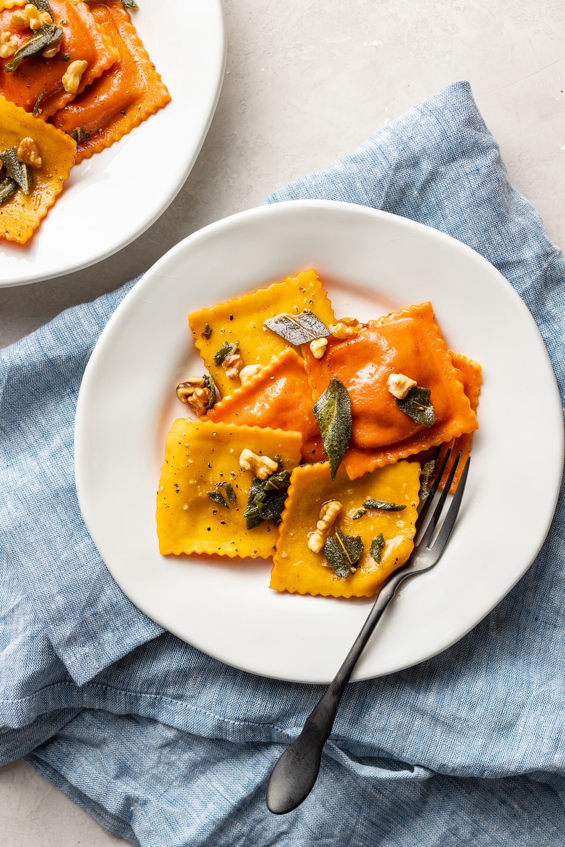 A plate with orange and yellow pumpkin ravioli garnished with sage brown butter sauce and toasted walnuts.