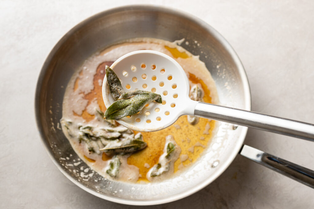 A white slotted spoon lifting crisped sage leaves out of the skillet.