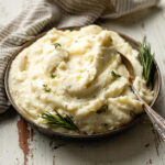 A bowl of fluffy garlic and herb mashed potatoes garnished with fresh rosemary and melted butter.