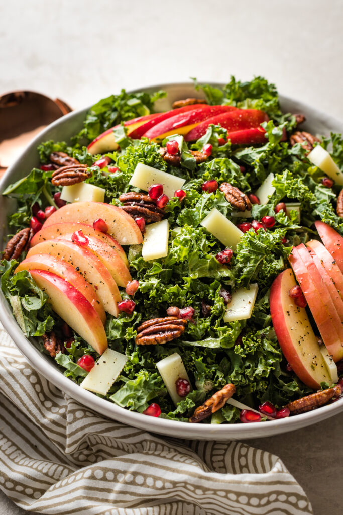 A large bowl of salad with kale, apples, Pecorino cheese, pomegranate seeds, and glazed pecans.