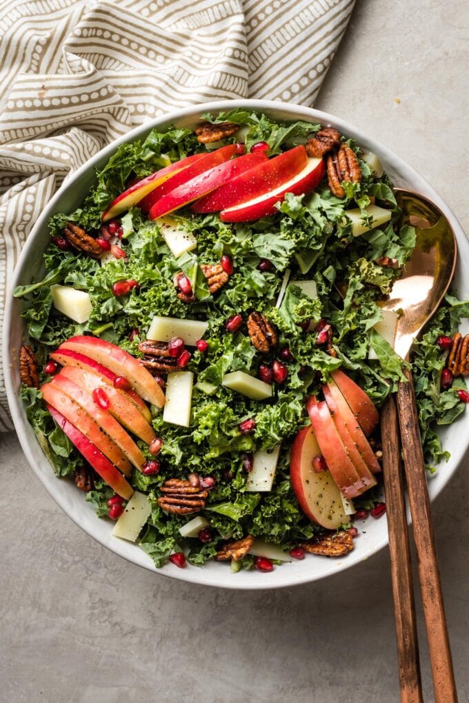 Salad bowl with a kale apple combination tossed with cheese, pecans, and pomegranate, with bronze salad tongs for serving.
