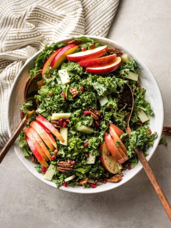 A large bowl of salad with kale, apples, Pecorino cheese, pomegranate seeds, and glazed pecans.