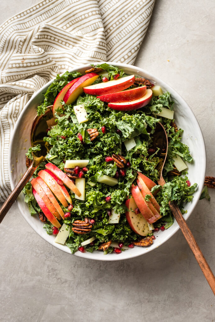 A large bowl of salad with kale, apples, Pecorino cheese, pomegranate seeds, and glazed pecans.