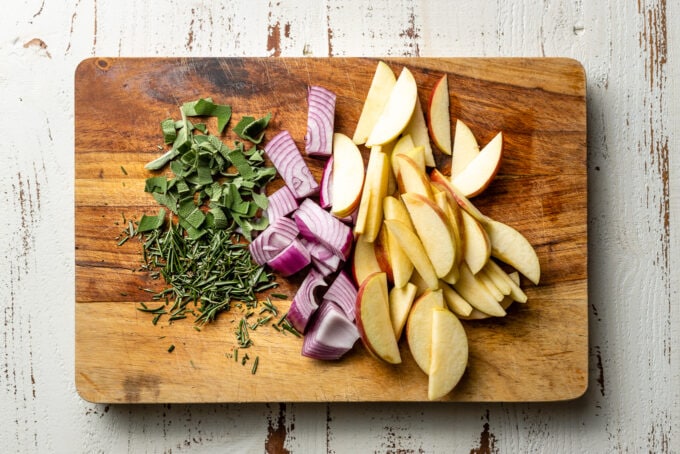 Chopped fresh rosemary and sage, chopped red onion, and chopped apples on a cutting board.