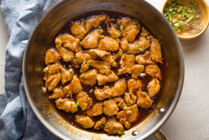 Skillet with bourbon chicken garnished with sliced green onions and toasted sesame seeds.