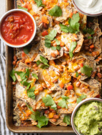 Baking sheet filled with pinto bean nachos, served with sour cream, guacamole, and salsa.