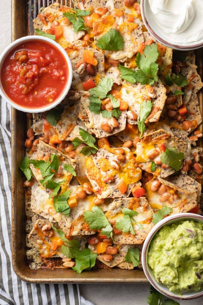 Baking sheet filled with pinto bean nachos, served with sour cream, guacamole, and salsa.