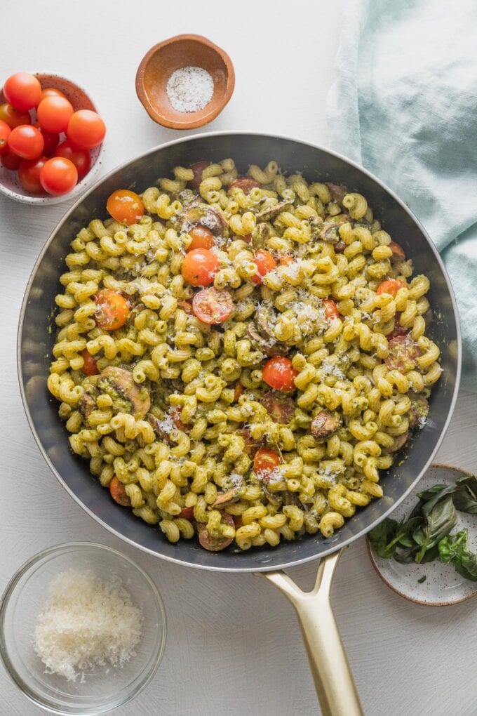 Kitchen counter with a skillet full of homemade pesto cavatappi with extra cherry tomatoes, Parmesan, and basil in the background.