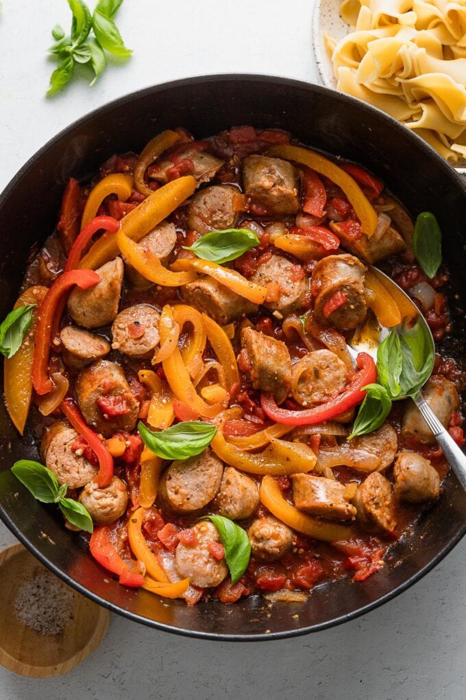 Overhead image of saucy Italian sausage and peppers with tomatoes and garlic in a deep-sided all-black cast iron skillet.