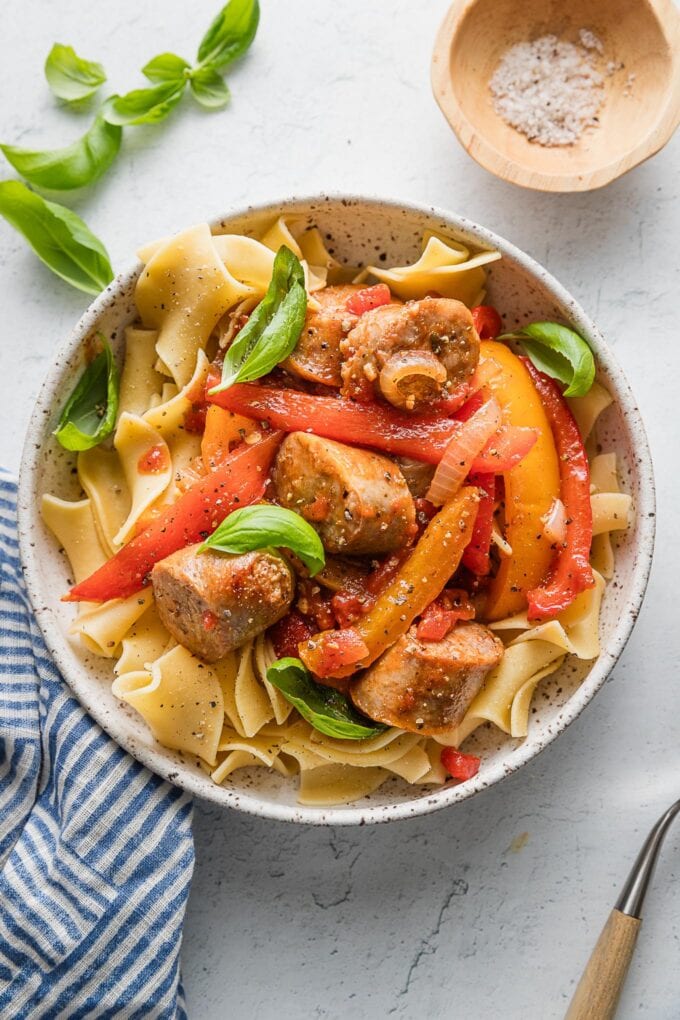 Countertop set with a bowl of Italian sausage, peppers, and onions in a light tomato garlic sauce, with fresh basil and extra sea salt nearby for sprinkling and seasoning to taste.