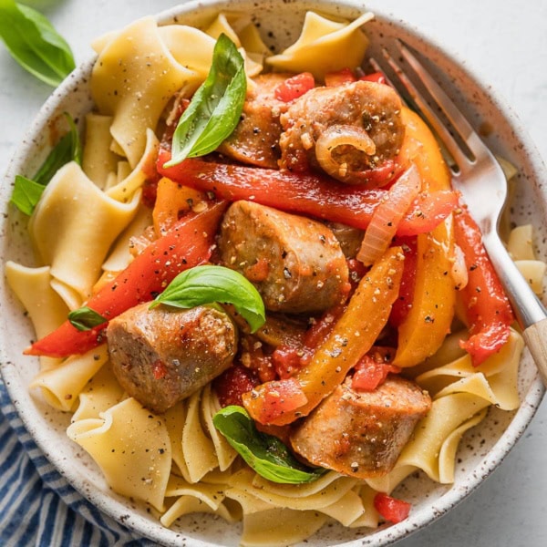 Bowl full of a one skillet saucy Italian sausage with peppers and onions served over egg noodles with fresh basil to garnish.