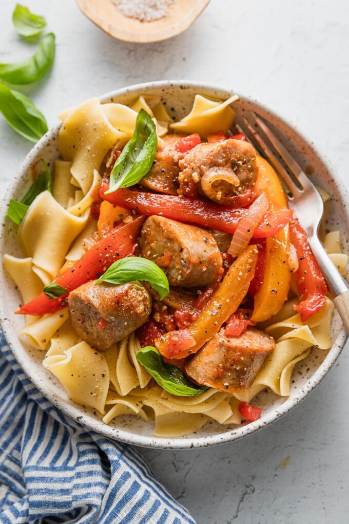 Bowl full of a one skillet saucy Italian sausage with peppers and onions served over egg noodles with fresh basil to garnish.