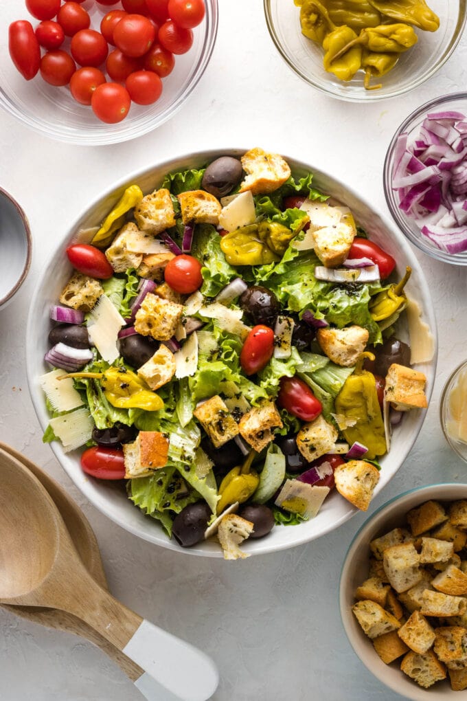 Bowl of salad surrounded by extra veggies and salad tongs ready to serve.