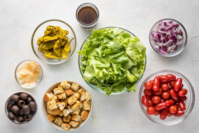 Prep bowls filled with washed romaine lettuce, Pepperoncini peppers, red onions, tomatoes, cheese, black olives, and croutons.