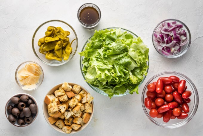 Prep bowls filled with washed romaine lettuce, Pepperoncini peppers, red onions, tomatoes, cheese, black olives, and croutons.