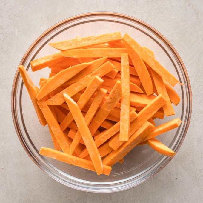 Three sweet potatoes cut into french fries, in a clear glass prep bowl.