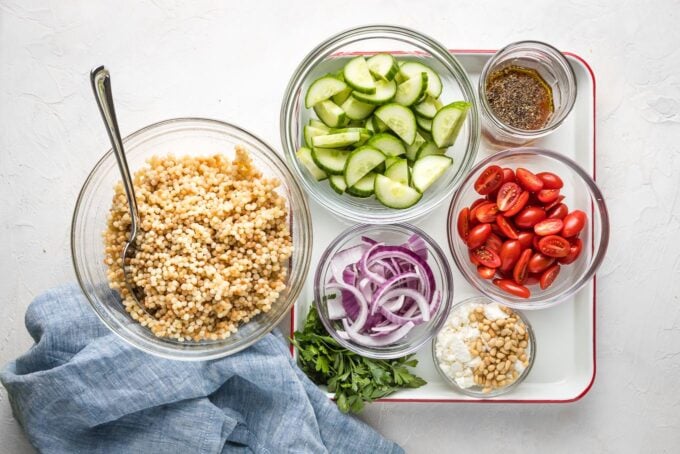 Pearl couscous, chopped cucumber, cherry tomatoes, red onion, feta, parsley, and salad dressing in small bowls.