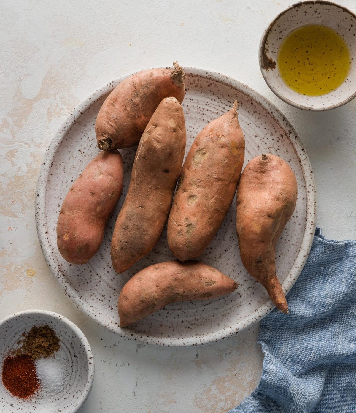 Whole sweet potatoes, olive oil, and a small bowl filled with chili powder, cumin, and kosher salt.
