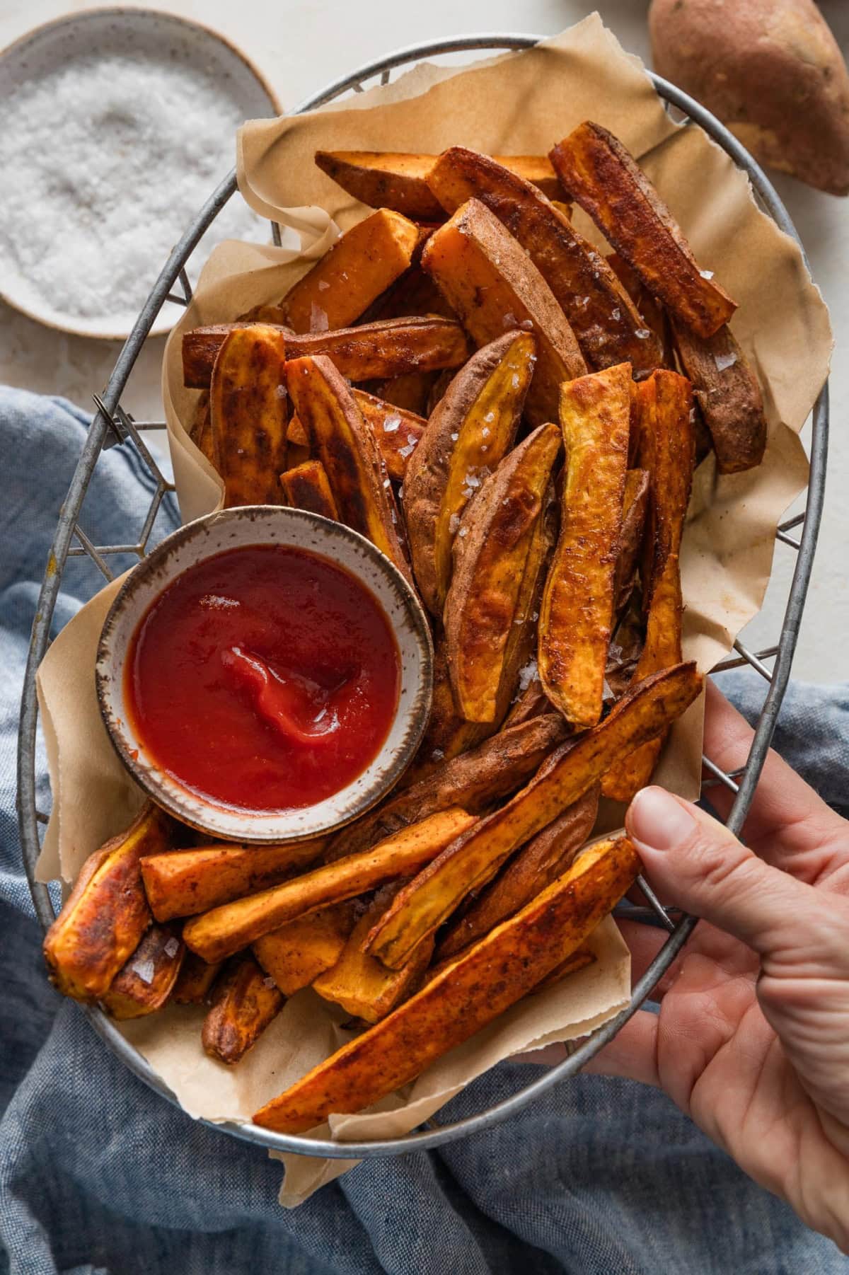 A woman's hand holding a metal basket lined with parchment paper and filled with oven-roasted homemade sweet potato wedges, topped with flaky sea salt and with a dipping bowl of ketchup.