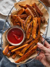 A woman's hand holding a metal basket lined with parchment paper and filled with oven-roasted homemade sweet potato wedges, topped with flaky sea salt and with a dipping bowl of ketchup.