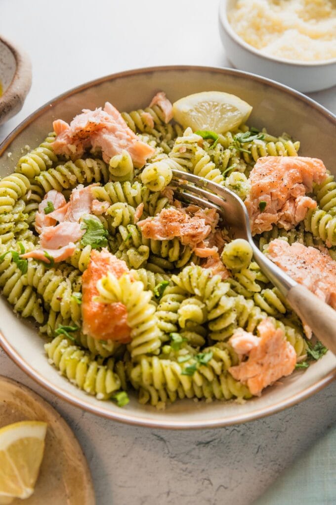 Angled view of a shallow bowl full of pesto pasta with roasted salmon, Parmesan, and lemon.