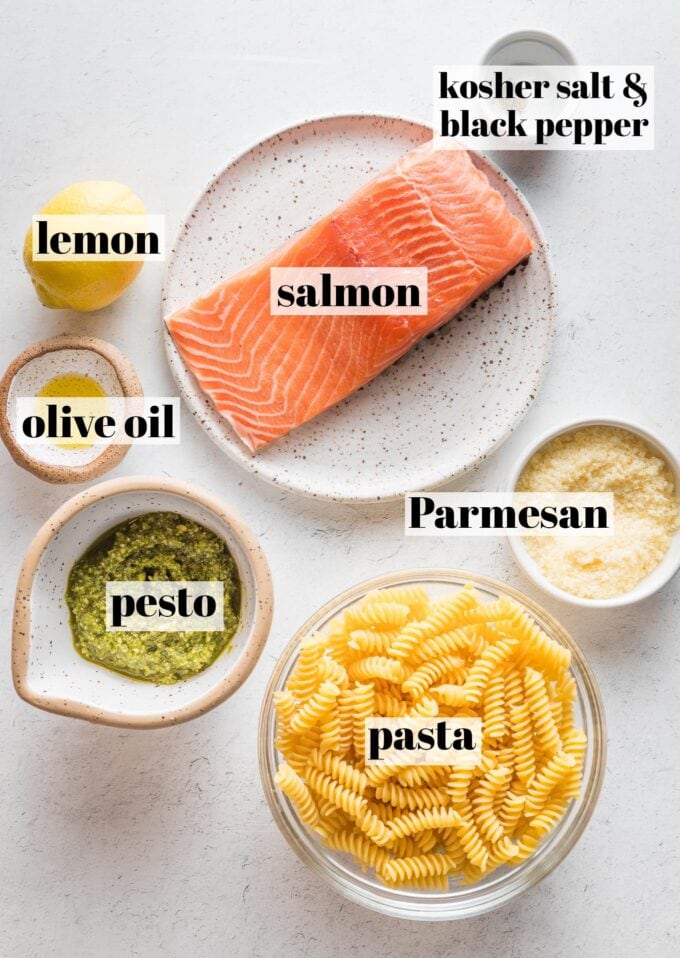 Labeled overhead photo of a salmon filet, dried pasta, basil pesto, a lemon, olive oil, Parmesan cheese, salt, and pepper all in prep bowls and ready to cook.