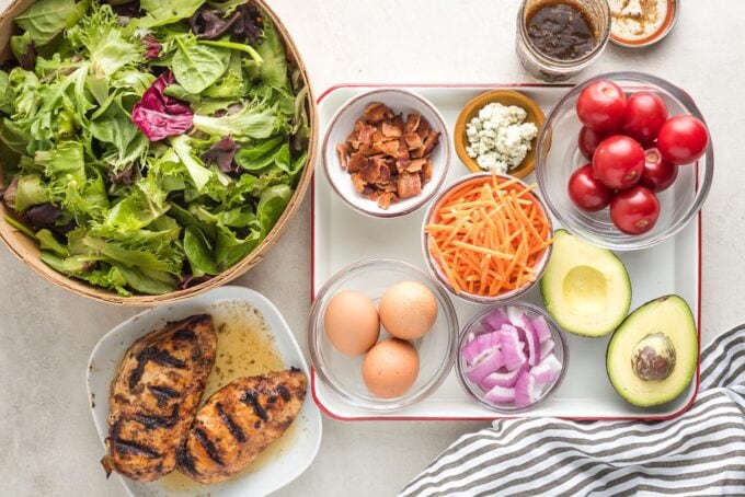 Prep bowls holding leafy greens, grilled balsamic chicken, cooked bacon, hard-boiled eggs, shredded carrots, blue cheese, chopped red onion, cherry tomatoes, and cut avocado.