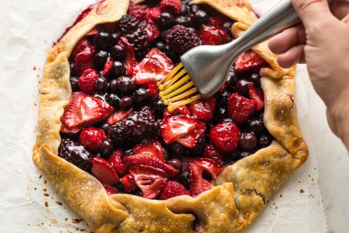 Close-up of a pastry brush brushing warm jam on top of a baked mixed berry galette.